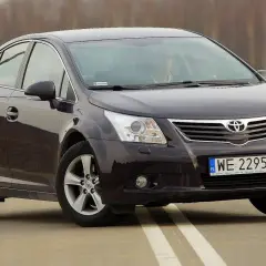 Ciemny sedan Toyota Avensis na drodze. Kierowca za kierownicą. Szczegółowe dane techniczne tego modelu są dostępne online.