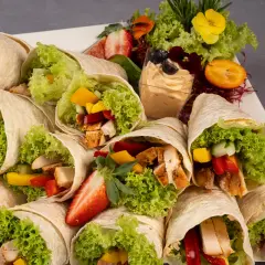 Mcwrap grillowany kurczak z warzywami i sałatą, ułożone na talerzu z dodatkami: dipem, owocami i ziołami.