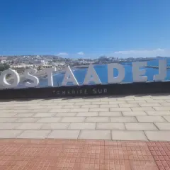 Napis "COSTA ADEJE TENERIFE SUR" na tle oceanu i miasta. Uważaj na słońce i tłumy, by w pełni cieszyć się wakacjami na Teneryfie.