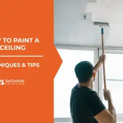 Learn how to paint a ceiling with this guide. A man uses a roller on an extension pole to apply white paint to a ceiling.
