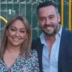 Kike Calleja y su pareja sonríen a la cámara. Ella lleva un vestido floral y él un traje azul.