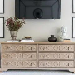 Ideas on how to decorate a dresser: a light wood dresser with decorative carvings, topped with books, a vase of flowers, a bust, and a dark vase.