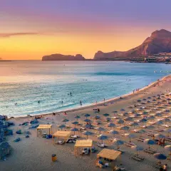 Ein langer Sandstrand mit vielen Liegestühlen und Sonnenschirmen. Das Meer ist ruhig, im Hintergrund Berge. Perfekt für die Frage: wo ist es im Mai warm Europa?