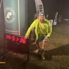 Uśmiechnięty biegacz z numerem 42 przy zegarze na trasie backyard ultra. Czołówka oświetla mu drogę.