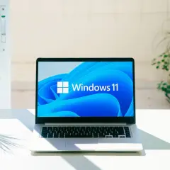Laptop z logo Windows 11, gotowy na aktualizację 25H2. Na biurku obok rośliny, w jasnym pomieszczeniu.