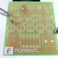 Płytka drukowana z elementami elektronicznymi, w tym rezystorami i diodami LED. Krokodylki podtrzymują płytkę, gotową do lutowania. Widać napis FORBOT.PL, sugerujący projekt z użyciem **tinol**u.