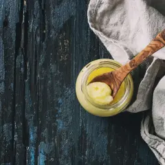 Ein Glas mit goldgelbem Ghee, daneben ein Holzlöffel mit einer Portion. Ghee ist geklärte Butter, ein vielseitiges Kochfett.
