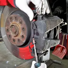 Mechanik w białych rękawiczkach dokonuje wymiany klocków hamulcowych, używając śrubokręta do podważenia starego klocka.