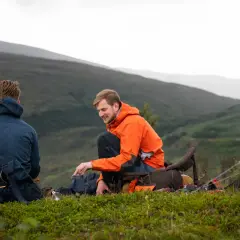 Två män njuter av en paus under vinter camping i en bergig miljö. En grön tält står uppsatt bredvid dem.
