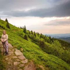 Karpacz-Śnieżka czas wejścia. Kobieta na kamienistym szlaku, zielone zbocza Karkonoszy, chmury na niebie.