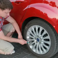 Mężczyzna w kratę sprawdza ciśnienie w kole czerwonego Forda. Wkrótce ruszy w dalszą podróż, tankując **benzynę**.