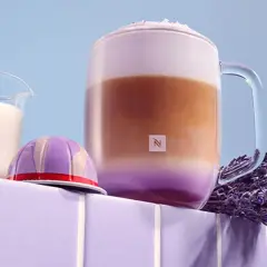 Ein aromatischer Lavendel Latte in einem Glasbecher, daneben eine Kaffeekapsel und Milch.