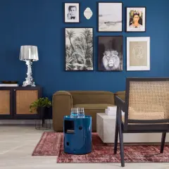 Ein stilvolles Wohnzimmer mit einer tiefblauen Wand, die mit Kunstwerken geschmückt ist. Ein Rattan-Stuhl steht vor einem Sofa, daneben ein runder, blauer Beistelltisch.