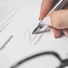 Dłoń z paznokciami malowanymi na różowo podpisuje dokument piórem. Widoczny napis "Signatures" i "Photographer's Signature". To ważny moment, jak wypowiedzenie po angielsku.