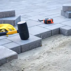 Młotek, rękawice i miarka czekają na ułożenie kostki. W tle widać przygotowany pod nią piasek, a nie beton.