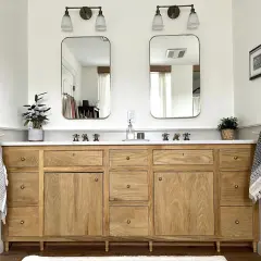 A beautiful DIY bathroom vanity with a natural wood finish, marble countertop, and brass fixtures. Two mirrors hang above, reflecting the bright, airy space.
