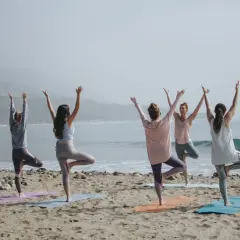 Cinco mujeres practican poses de yoga para 4 en la playa, con los brazos levantados hacia el cielo.
