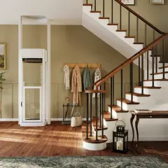 Ein moderner Treppenlift mit Glaskabine steht neben einer Holztreppe. Bilder und Garderobe schmücken die Wand.