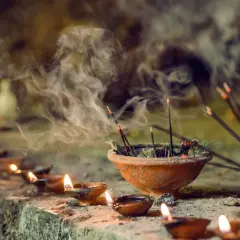 Humo de incienso llenando el aire, un recordatorio de que el incienso es malo para los pulmones. Velas encendidas y estatuillas borrosas en el fondo.