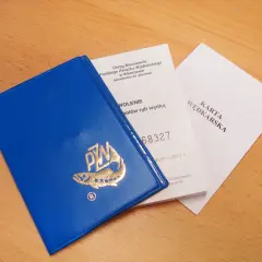 Niebieska książeczka z logo PZW i dokumenty "Karta Wędkarska" na drewnianym tle.