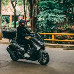 Mujer con casco y ropa negra conduce una moto Yamaha sin marchas, con caja trasera, por una calle arbolada.