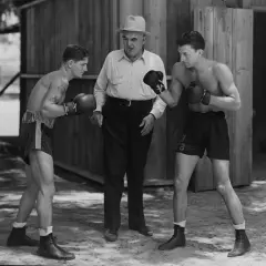 Two boxers spar while a trainer watches. The boxer on the left has a tattoo. This scene evokes the spirit of legendary boxer James J. Jeffries.