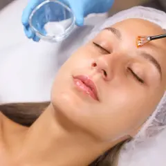 Manos enguantadas aplican un peeling facial con brocha a una mujer con los ojos cerrados.