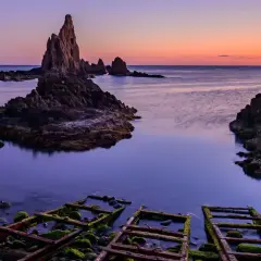 Rocas afiladas emergen del mar sereno bajo un cielo crepuscular. La **sierra Cabo de Gata** se revela en su esplendor, con restos oxidados de estructuras en primer plano.