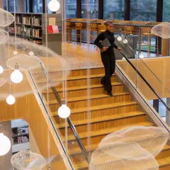Kobieta z laptopem schodzi po drewnianych schodach w nowoczesnej bibliotece. Zastanawia się, ile trwają studia, gdy mija wiszące dekoracje w kształcie chmur.