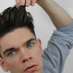 Mann mit einem messy quiff, der sich die Haare hochzieht. Blaue Augen, graues Sweatshirt.