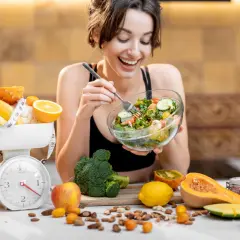 Mujer sonriente come ensalada. Alrededor, frutas, verduras y una báscula, sugiriendo una tabla de dieta para engordar.
