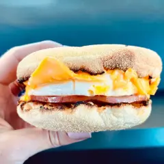 Mcmuffin farmerski z jajkiem sadzonym, plastrem szynki i roztopionym serem, trzymany w dłoni.