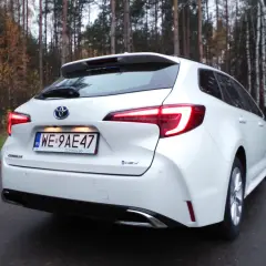 Biała Toyota Corolla 1.8 Hybrid na tle jesiennego lasu. Widok z tyłu, z podświetlonymi światłami.