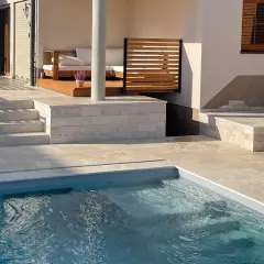 Piscina con bordo in pietra travertine. Gradini e pavimentazione in travertine creano un'elegante area relax con divani in legno.