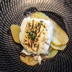Bacalao gratinado con alioli, servido sobre patatas doradas en un plato negro texturizado.