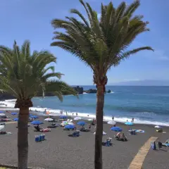 Palmy nad czarną plażą w Puerto de Santiago, gdzie turyści odpoczywają pod parasolami.