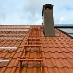 Pomarańczowe stopnie kominiarskie do dachówki prowadzą do komina na dachu z panelami fotowoltaicznymi.