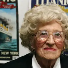 Una anciana con gafas grandes sonríe. Detrás de ella, carteles del Titanic, evocando a los supervivientes del trágico viaje.