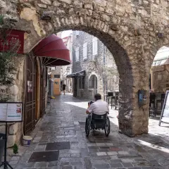 Wąska, brukowana uliczka w **Budva stare miasto**. Mężczyzna na wózku inwalidzkim przejeżdża pod kamiennym łukiem. Po bokach sklepy i restauracje.