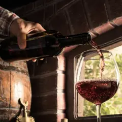 Ręka nalewa wino z czerwonego winogrona do kieliszka. Obok drewniana beczka i ceglana ściana.