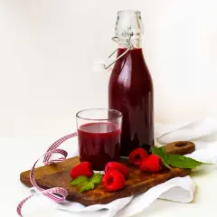 Szklanka i butelka z sokiem malinowym, maliny i listki mięty na desce.