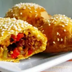 Złociste empanadas posypane sezamem, z widocznym farszem z warzyw i mięsa. Pyszna przekąska.