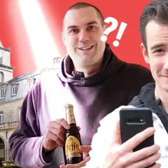 Deux hommes souriants devant un bâtiment historique, l'un tenant une bouteille de Leffe.