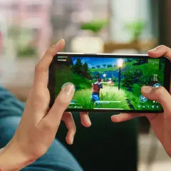 Ręce trzymają smartfon, na którym widać fragment popularnej gry na telefon. W tle widać nogi w jeansach i trampkach.