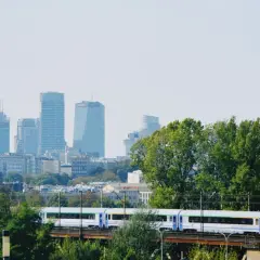 Pociąg PKP jedzie przez zielone drzewa z widokiem na warszawskie wieżowce. Szukasz tanich biletów PKP Warszawa - Kraków?
