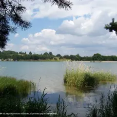 Spokojny zalew Dźibice otoczony zielenią, z kempingiem na brzegu i chmurami na niebie.