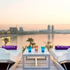 Dos tumbonas blancas con toallas moradas de W Hotels, una mesa con vasos y velas, y vistas al mar y la ciudad de Barcelona al atardecer.