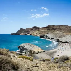 Vista de una playa con gente disfrutando del sol y el mar azul, con formaciones rocosas y montañas al fondo. A lo lejos se divisa el faro de los genoveses.