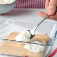 Nakładanie łyżką puszystego kremu z mleka skondensowanego i mascarpone na biszkopty w szklanej formie.