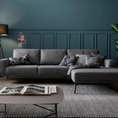 Ein moderner Wohnbereich mit einem grauen Ecksofa, das auf einem gemusterten Teppich steht. Ein Couchtisch mit einem offenen Buch und eine Stehlampe vervollständigen das Bild.